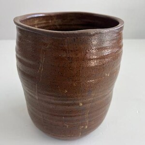 Puede incluir: Un vaso de cerámica marrón hecho a mano con una superficie texturizada y acanalada. La taza tiene una abertura ancha y un borde ligeramente irregular. El exterior es de un rico color marrón terroso, con sutiles variaciones de tono.