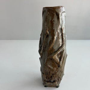 Op de afbeelding: Een hoge, cilindrische keramische vaas met een getextureerd, onregelmatig oppervlak. De buitenkant heeft een gevlekt bruin en grijs glazuur met diepe, hoekige inkepingen en verhoogde ribbels die een robuust en organisch uiterlijk creëren.