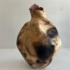 Puede incluir: Un recipiente de madera tallado a mano con una forma orgánica e irregular. La superficie presenta una veta de madera de color tostado claro contrastada por marcas negras carbonizadas y un interior oscuro y texturizado visible en la abertura.