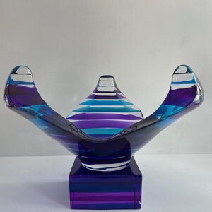 May include: A decorative glass bowl with a square base. The bowl is a vibrant mix of blue and purple hues, with horizontal stripes. The bowl's design features curved, wing-like sides and a central, conical element.