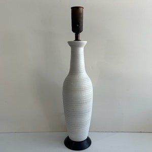 May include: A tall, white ceramic table lamp base with a slender, bottle-like silhouette and horizontal ribbed texture. The lamp features a dark brown metal socket at the top and rests on a circular, solid black base at the bottom.