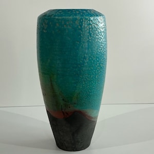 May include: A tall, turquoise ceramic vase with a textured surface. The vase has a dark gray base and a gradient of colors, including teal, green, and red. The vase is a decorative item.