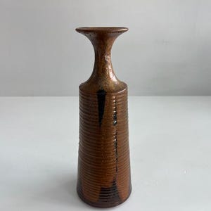 May include: A tall, cylindrical ceramic vase with a flared rim and a narrow neck. The body features a textured, ribbed surface with a glossy brown glaze, accented by darker brown vertical streaks and a mottled finish throughout the exterior.