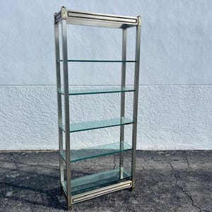 May include: A tall, silver-toned metal and glass shelf unit with four glass shelves. The shelves are rectangular and appear to be approximately 12 inches deep. The frame has decorative gold accents at the top and bottom. The unit stands against a white textured wall.