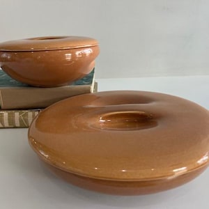 May include: Two round, terracotta-colored ceramic bowls with matching lids. Each bowl features a smooth, glossy finish and a central indentation on the lid. The bowls are positioned near two stacked books with visible text on their spines.