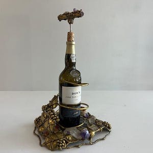 May include: A gold-toned metal wine bottle holder featuring a decorative base with sculpted grape leaves and clusters, accented with purple amethyst stones. A bottle of Dow's Fine Tawny Port is held in the center, topped with a matching corkscrew.
