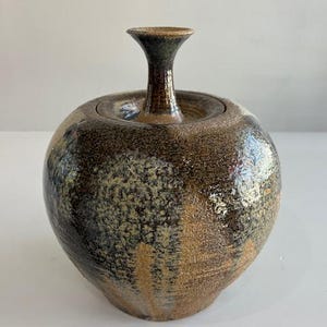 May include: A handcrafted ceramic jar with a lid and a slender, flared neck. The jar features a speckled, earthy color palette of brown, tan, and gray, with a glossy finish. This decorative piece is suitable for home decor.