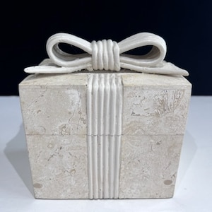 May include: A square box made of light beige, speckled stone with a natural textured finish. The box features a carved stone ribbon and bow detail on the lid and front, mimicking a wrapped gift. The stone has visible natural mineral variations.