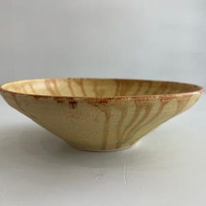 Puede incluir: Un cuenco de cerámica poco profundo con un borde ancho y acampanado. El exterior y el interior presentan un esmalte beige arena texturizado con vetas verticales y goteantes de pigmento marrón rojizo que se extienden desde el borde hasta la base.