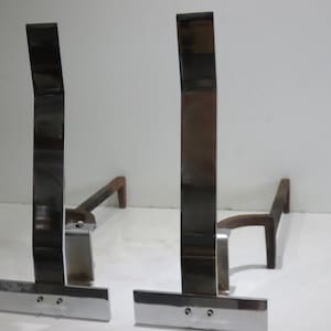 Puede incluir: Un par de atriles para chimenea. Cada atril tiene un montante alto, pulido, rectangular, de metal oscuro, una barra horizontal y un soporte corto e inclinado. La base es de metal brillante color plata.