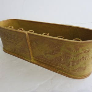 Peut inclure: Un bol à fleurs ou centre de table en céramique de forme ovale allongée avec un vernis jaune moutarde. L'extérieur présente un motif géométrique répétitif texturé et l'intérieur contient une série de petites ouvertures circulaires.