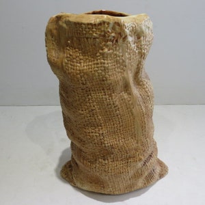 May include: A tan ceramic vase shaped like a standing burlap sack. The exterior features a highly detailed, raised woven texture that mimics the appearance of coarse fabric, with a wide, open top and a slightly flared, uneven base.