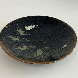 Puede incluir: Un plato pequeño y redondo de cerámica con un fondo oscuro moteado. El plato presenta un diseño pintado a mano de hojas verdes y bayas blancas. El borde del plato tiene un tono marrón terroso.