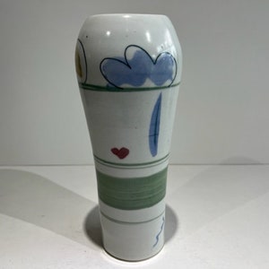 May include: A white ceramic vase with a tapered cylindrical shape, featuring hand-painted motifs including a blue cloud, a small red heart, a vertical blue dash, a wavy blue line, and horizontal green bands encircling the lower section.