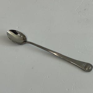 May include: A stainless steel spoon with a rounded bowl and a long, slender handle. The handle has a circular design at the end. The spoon is set against a white background.