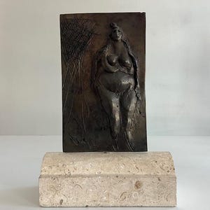 May include: A bronze relief sculpture of a woman on a rectangular plaque, mounted on a light-coloured stone base. The sculpture depicts a full-figured woman with long hair. The background features abstract lines. The base is rectangular with rounded edges.