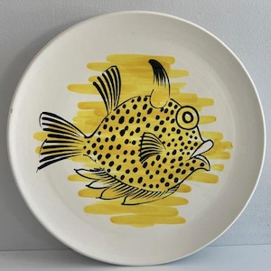 May include: A white ceramic plate with a quirky fish illustration. The fish is yellow with black spots and outlines, set against a yellow backdrop. The plate has a simple, clean design, suitable for serving or display.
