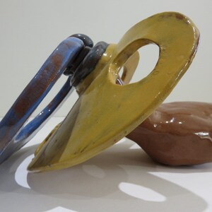 Puede incluir: Escultura abstracta de cerámica con una combinación de colores azul, amarillo y marrón. La escultura presenta un elemento circular azul, una pieza amarilla con un agujero y una forma redondeada marrón. La obra está sobre un fondo blanco.