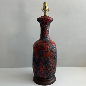 Puede incluir: Lámpara de mesa alta de cerámica texturizada en rojo, negro y marrón. La lámpara tiene forma de botella con cuello estrecho y base ancha. Se apoya en una base de madera marrón oscuro y tiene un portalámparas de latón. Aproximadamente 40 cm de altura.