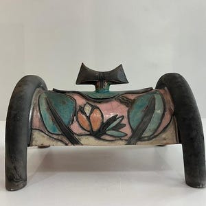Puede incluir: Una caja de cerámica con tapa curva y un asa de acabado mate oscuro en forma de lazo. El cuerpo presenta un motivo floral pintado a mano en tonos rosa suave, azul verdoso y crema, enmarcado por patas arqueadas oscuras y texturizadas.