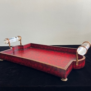 May include: A rectangular decorative tray with a red mottled finish and bronze-colored trim. It features clear, cylindrical handles and small, round feet. The tray is designed for serving or display.