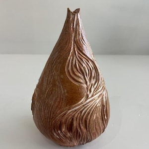 Puede incluir: Un jarrón marrón en forma de lágrima con una superficie texturizada y una abertura estrecha. El jarrón presenta un diseño tallado y fluido en un color marrón más claro, agregando interés visual. Es un artículo decorativo.