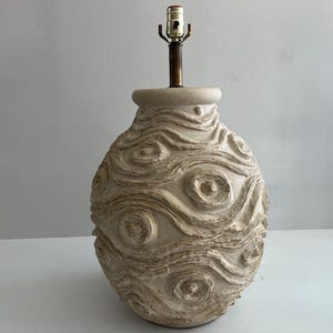 May include: A beige ceramic table lamp base with a rounded, bulbous shape featuring a raised, swirling, textured pattern across the surface. The lamp includes a central brass-colored metal socket assembly extending from the top opening.