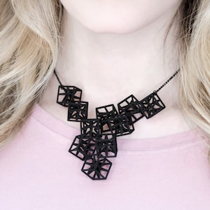 Può includere: Una collana geometrica nera con una serie di forme a cubo interconnesse. La collana è indossata da una persona con lunghi capelli biondi.