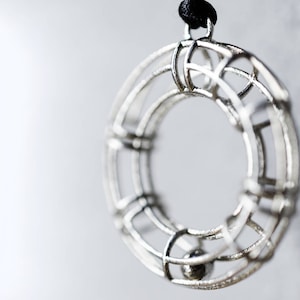 Pode incluir: Um colar com um pingente de prata de design circular e geométrico. O pingente é feito de múltiplos anéis interconectados, criando um efeito de gaiola. Uma pequena bola de prata pendura dentro da gaiola.
