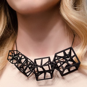 Può includere: Una donna con lunghi capelli biondi indossa una collana geometrica nera. La collana è composta da diversi cubi interconnessi.