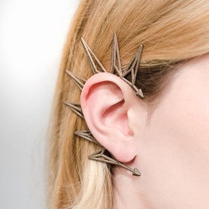 Puede incluir: Primer plano de la oreja de una mujer con un pendiente de oreja de plata. El pendiente de oreja está hecho de múltiples formas triangulares y tiene un diseño puntiagudo.