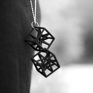 Op de afbeelding: Een zilveren ketting met een zwarte geometrische hanger gemaakt van twee in elkaar grijpende kubussen.