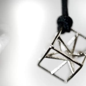 Op de afbeelding: Een zilveren geometrische hanger in de vorm van een kubus met een zwart koord.