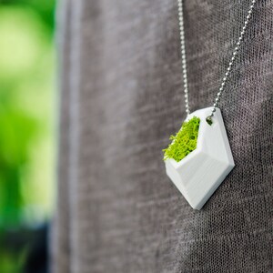 May include: A white geometric pendant necklace filled with bright green moss. The pendant is diamond-shaped and hangs from a silver ball chain. The background is a blurred green and gray.