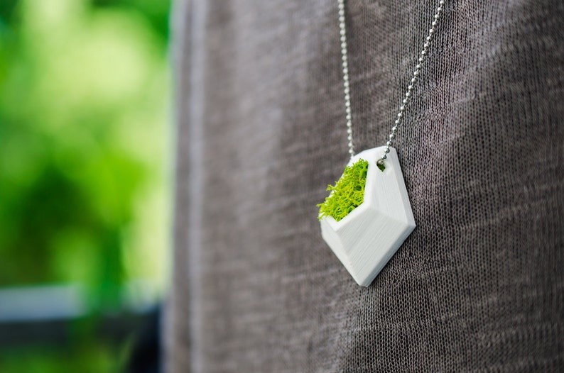 May include: A white geometric pendant necklace filled with bright green moss. The pendant is diamond-shaped and hangs from a silver ball chain. The background is a blurred green and gray.