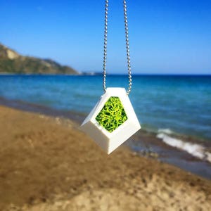 May include: A white geometric pendant necklace with a silver chain. The pendant has a diamond shape with a green moss filling. The background shows a beach with blue water and sky.