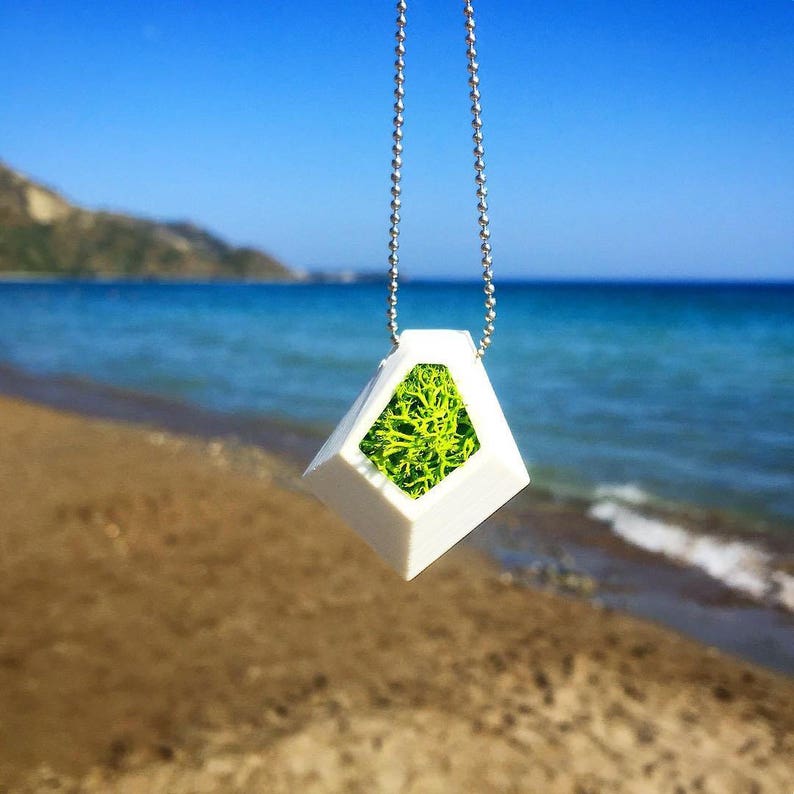 May include: A white geometric pendant necklace with a silver chain. The pendant has a diamond shape with a green moss filling. The background shows a beach with blue water and sky.