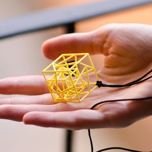 Può includere: Una collana con un ciondolo geometrico giallo e un cordino nero. Il ciondolo è un design a forma di cubo 3D con un motivo complesso e intrecciato.