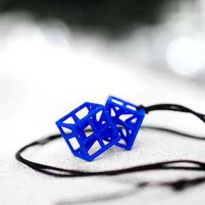 Könnte beinhalten: Eine blaue geometrische Halskette mit zwei würfelförmigen Anhängern. Die Halskette besteht aus einem schwarzen Band.