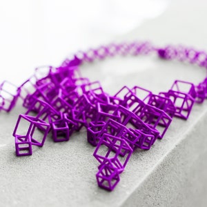 Può includere: Una collana viola fatta di forme di cubi aperti interconnessi. La collana è su una superficie grigia.