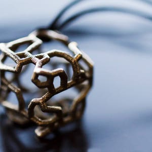 Op de afbeelding: Een gouden metalen ketting met een geometrische, opengewerkte hanger. De hanger heeft de vorm van een kubus met ingewikkelde, met elkaar verbonden lijnen.
