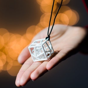 Może przedstawiać: Naszyjnik z białym, geometrycznym, sześciennym zawieszką i czarnym sznurkiem. Zawieszka wykonana jest z delikatnego, skomplikowanego wzoru z drutu.