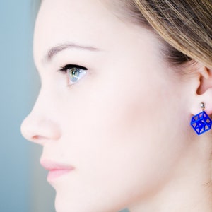 Op de afbeelding: Een vrouw draagt ​​een paar blauwe geometrische oorbellen. De oorbellen zijn kubusvormig met een draadframe-ontwerp.