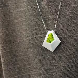 May include: A white geometric pendant necklace with a silver chain. The pendant has a diamond shape with a hollow center filled with bright green moss. The necklace is displayed against a gray fabric background.