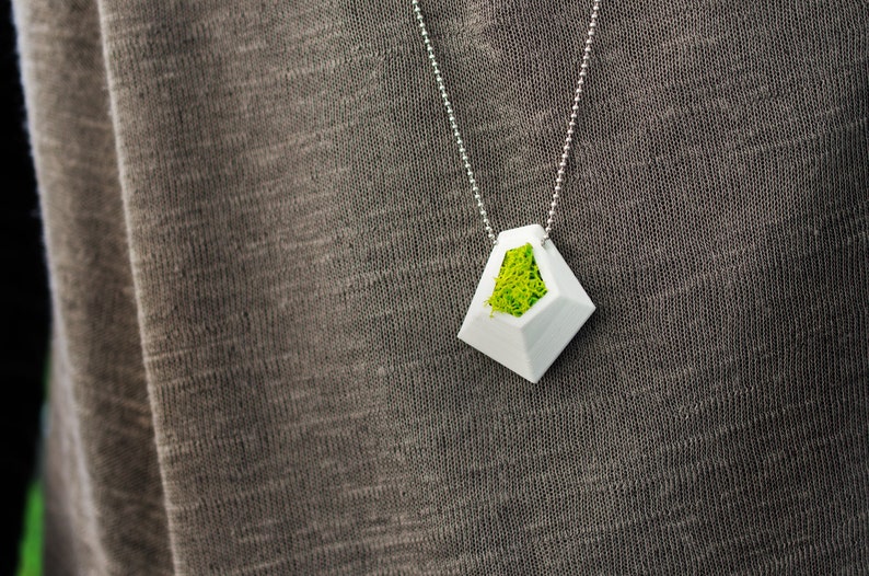 May include: A white geometric pendant necklace with a silver chain. The pendant has a diamond shape with a hollow center filled with bright green moss. The necklace is displayed against a gray fabric background.