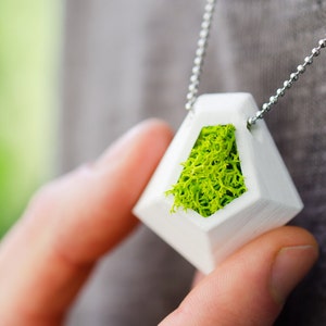 May include: White geometric pendant necklace with a bright green moss filling. The pendant hangs from a silver ball chain. The necklace is held by a person.
