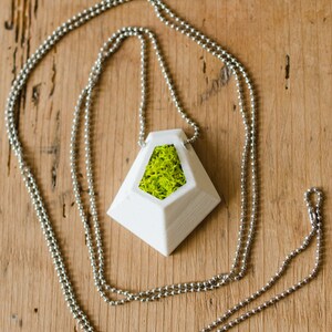 May include: A white geometric pendant necklace with a silver chain. The pendant has a triangular shape with a hollow center filled with bright green moss. The necklace is displayed on a wooden surface.