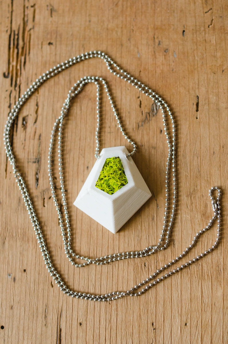 May include: A white geometric pendant necklace with a silver chain. The pendant has a triangular shape with a hollow center filled with bright green moss. The necklace is displayed on a wooden surface.
