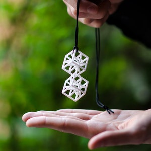 Op de afbeelding: Een witte geometrische ketting met twee gestapelde, opengewerkte kubusvormen die aan een zwart koord hangen.