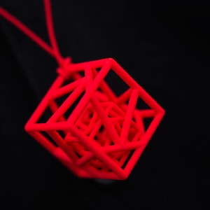 Può includere: Un ciondolo geometrico rosso brillante stampato in 3D a forma di cubo. Il cubo è composto da più strati di linee che si intersecano, creando un design complesso e intricato.